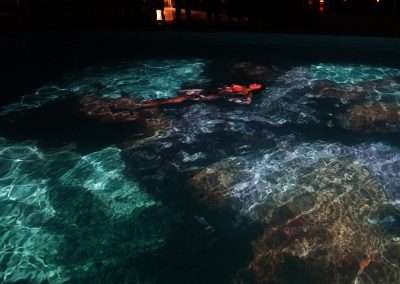 A person floats on their back in a dimly-lit indoor pool at Bishop's Lodge, illuminated with blue and purple-tinted lights. The surface of the water reflects the lighting, creating a serene atmosphere reminiscent of an Eric Tillinghast installation. The scene conveys a sense of calm and tranquility.