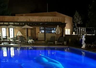 A nighttime scene at Bishop's Lodge shows a person floating in a well-lit pool, surrounded by lounge chairs, umbrellas, and a building with illuminated windows in the background. The water glows blue, creating an eerie yet serene atmosphere reminiscent of Eric Tillinghast's evocative art installations.