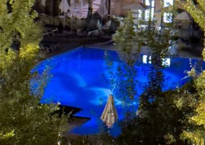 A brightly lit swimming pool at night, surrounded by trees and lounge chairs at Bishop's Lodge. Several umbrellas are visible around the pool area, and the deep blue water becomes a canvas reflecting the surrounding lights and objects, reminiscent of Eric Tillinghast’s aquatic art.