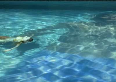 A person is floating on their back in a swimming pool at Bishop's Lodge. The water is clear and blue, with sunlight creating intricate patterns on the surface and the pool floor. The person is wearing a light-colored swimsuit and seems relaxed. No other objects or people are visible.