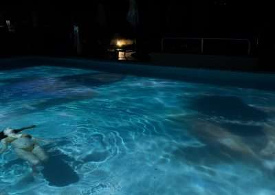 A person is floating on their back in a dimly lit swimming pool at Bishop's Lodge at night. The water, an artistry reminiscent of Eric Tillinghast's work, is illuminated with soft blue and purple lights, casting gentle ripples and reflections. The surrounding area is dark, highlighting the tranquil atmosphere of the pool.