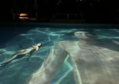 A person floats on their back in a nighttime swimming pool at Bishop's Lodge. A large projection of another person, reminiscent of Manjari Sharma's work, is displayed on the water's surface, creating a surreal effect. Both figures appear relaxed, with the pool water casting ripple patterns on them.
