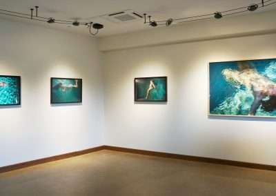 A gallery room with four framed photos on a wall, each depicting underwater scenes with human figures swimming. Three smaller photos are aligned on the left side, while a larger photo is displayed toward the right. The room is lit by spotlights.