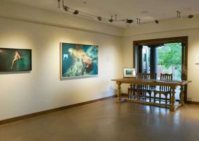 A spacious art gallery room with beige walls featuring two underwater-themed paintings. A large wooden table with chairs sits near an open doorway leading outside. The gallery is lit with track lighting, highlighting the artwork on display.