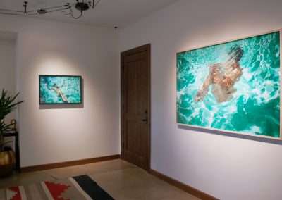 A gallery space with white walls displays two framed paintings of swimmers diving into turquoise water. There is a wooden door between the artworks, a plant in a pot on the left, and a geometric-patterned rug on the floor. The area is softly lit.