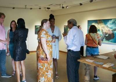 People are gathered in an art gallery, engaging in conversation and viewing artwork on the walls. Some are holding drinks while others are observing the exhibits. Various pieces of art are displayed in the background. The atmosphere appears social and relaxed.