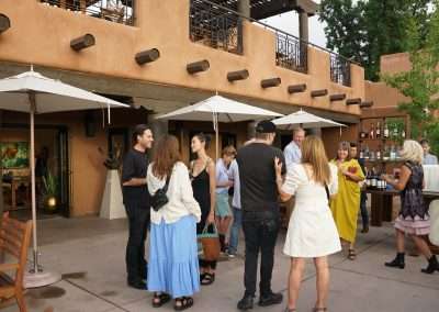 A group of people are gathered on an outdoor patio, conversing and enjoying the event. They stand near large umbrellas and a building with a rustic adobe design. Several tables are set up with drinks, and there are trees in the background.