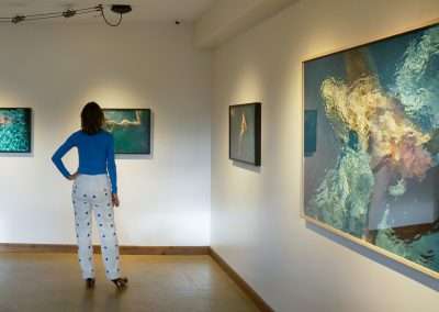 A person in a blue top and white pants with patterns stands in an art gallery, observing photographs on the walls. The photographs depict underwater scenes with human figures, featuring shades of blue and green. The gallery has a minimalist design.