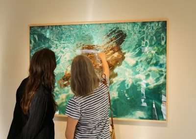 Two individuals stand before a framed painting of a person swimming underwater. The person on the right points to a specific detail on the artwork, while the one on the left observes. The gallery setting has neutral walls, focusing attention on the vibrant painting.
