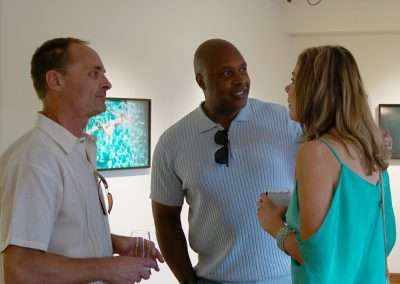 Three people are engaged in conversation at an art gallery. The man on the left holds a wine glass, the man in the middle is wearing a light blue shirt with sunglasses, and the woman on the right is dressed in a teal top. They are standing in front of framed photographs on the wall.