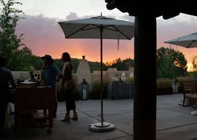 People conversing near a mobile food cart on a patio beneath large umbrellas at sunset. The clear sky is tinged with hues of orange and purple. Green trees and various outdoor seating and decor are visible in the background.