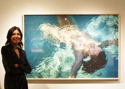 A person stands in front of a large framed photograph of a figure underwater. The water's surface creates a rippled effect, distorting the figure. The person is smiling and has their arms crossed.