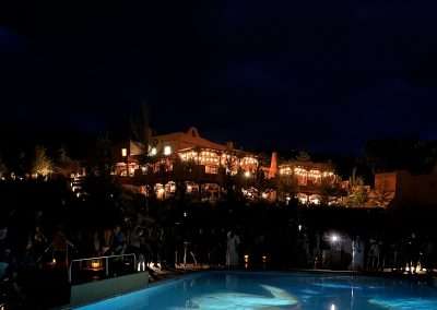 A brightly lit building casts warm light against the night sky, reflected in a nearby swimming pool that is illuminated from below. A crowd gathers near the poolside, creating a serene and enchanting evening atmosphere.