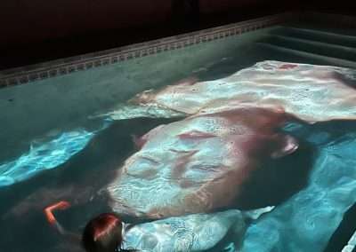 A person floating face-up in a swimming pool with vivid light patterns projected on the water's surface. A second person, reminiscent of Manjari Sharma's ethereal photography, stands by the edge of the pool, partially visible and capturing the scene. The setting is dark with dramatic lighting effects.