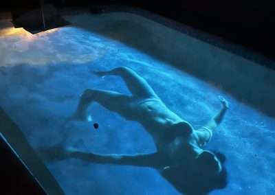 A person is floating on their back in an illuminated swimming pool at night. The pool water emits a blue glow, casting a serene and eerie ambiance. The scene is quiet and atmospheric, reminiscent of Manjari Sharma's evocative photographic projection work.