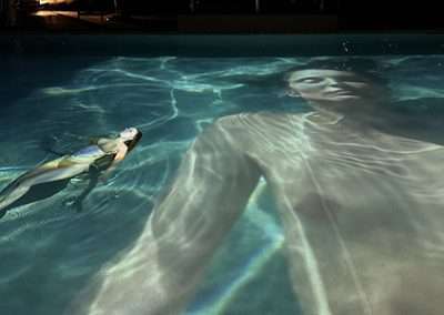 A person is floating on their back in a swimming pool at night. The water's surface displays a large, projected image of a woman in a white outfit. The projection creates an artistic and surreal effect, reminiscent of art exhibitions, blending the floating person with the image.