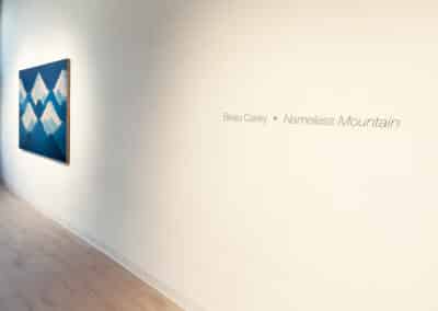 A minimalist art gallery room with light wooden floors. The left wall displays a painting of blue and white mountains. On the right wall, text reads "Beau Carey • Nameless Mountain" in simple fonts. The room is well-lit with natural light.
