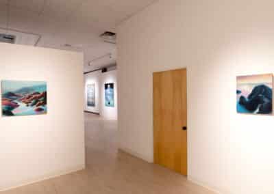 An art gallery with minimalist decor displays several framed landscape paintings on white walls. The paintings feature mountainous scenes in vibrant colors. A wooden door is visible on the right-hand wall, and the space is well-lit with soft lighting.