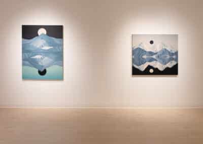 A minimalist art gallery with two paintings on a light beige wall. Both pieces feature mountainous landscapes with reflective water below and geometric shapes, one in darker shades of blue and the other in lighter tones. The gallery floor is light wood.