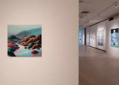 A spacious art gallery with white walls and light wooden flooring. On the left wall, a colorful painting of abstract mountains and water is displayed. The gallery extends into the background, showcasing several more paintings with similar abstract themes.