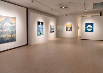 A modern art gallery featuring various paintings of mountainous landscapes in different color schemes. The room is well-lit with spotlights, and the walls and floor are white, giving a clean and minimalist look.