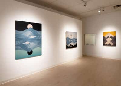A modern art gallery featuring minimalist landscape paintings on white walls. The artworks depict abstract mountain scenes with reflections, utilizing muted and calming colors. The gallery is well-lit, highlighting the paintings’ details and textures.