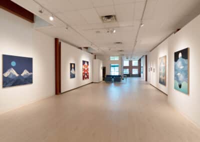 A spacious art gallery with white walls displaying various paintings, mainly landscape and abstract art. The floor is light wood, and the gallery is well-lit with ceiling lights. At the far end, a doorway and windows allow natural light to enter the room.