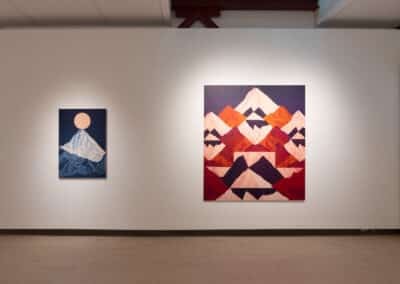 Two landscape paintings hang on a white gallery wall. The left painting depicts a snowy mountain under a circular sun. The right painting showcases multiple mountains in shades of red, orange, and purple. The room has a wooden floor and a lit ceiling.