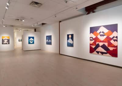 A modern art gallery showcasing abstract mountain paintings in various colors and styles. The walls are adorned with several pieces, including vibrant red and orange mountains, blue and white peaks, and others in the background. The gallery is well-lit and spacious.