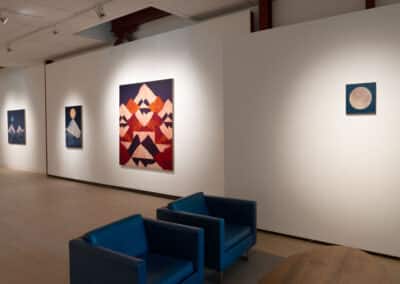 An art gallery featuring several paintings on white walls. The artwork includes geometric and abstract designs, with prominent pieces depicting mountainous shapes. Two blue chairs are placed in front of the artwork, creating a space for viewing and contemplation.
