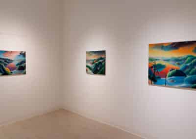 A clean, white-walled gallery displaying three vibrant landscape paintings, each featuring lush, colorful scenes with hills and rivers. The leftmost painting is smaller, while the right two are larger. The lighting highlights the vivid colors and details of the artwork.