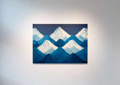 A painting depicting a series of blue and white mountain peaks on display in an art gallery with a plain, light-colored wall as the background. The highest peak is centrally placed and stands out in a lighter white shade among the other blue-toned peaks.
