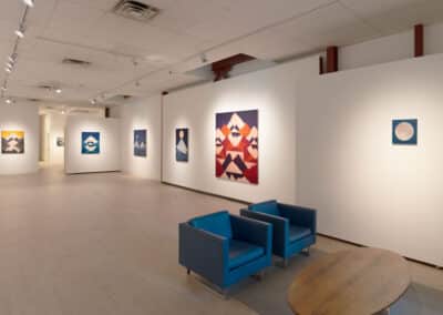 An art gallery features multiple abstract paintings with geometric designs on white walls. In the foreground, two blue armchairs and a wooden oval table are arranged. The room is spacious and well-lit by ceiling lights.