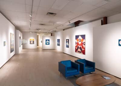 A spacious art gallery with white walls and light wood flooring displays modern, geometric paintings. There are two blue armchairs and a round wooden table in the foreground. The gallery is well-lit with track lighting.