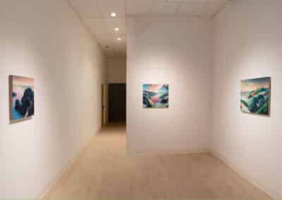 A narrow art gallery with white walls displays three landscape paintings featuring abstract, colorful terrain. Two paintings are hung on the side walls and one on the back wall. The room has a light wooden floor and recessed ceiling lights.