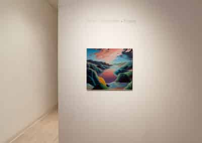 A painting titled "Edges" by Adam Sorensen is displayed on a gallery wall. The artwork features an abstract landscape with colorful hills and a reflective body of water under a sky with pink clouds. The setting appears to be a well-lit exhibition space.