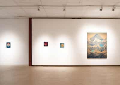 An art gallery featuring four framed artworks on a white wall. Three smaller circular pieces are on the left, each with varying colors and patterns. On the right, a larger painting depicts a mountain landscape with blue and beige tones. Overhead spotlights illuminate the artwork.