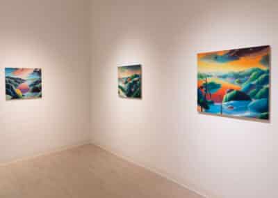 A gallery room with three vibrant landscape paintings on white walls. Each painting depicts colorful, abstract scenery with rolling hills, mountains, and bodies of water. The room has light wooden flooring and soft, even lighting illuminating each artwork.
