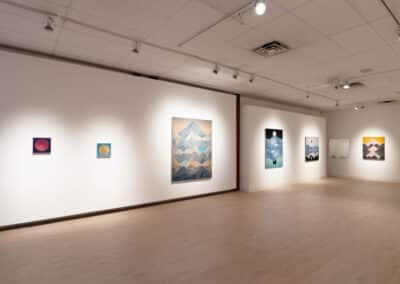 A contemporary art gallery with white walls and wooden floors. Various abstract paintings, including landscapes and geometric patterns, are displayed on the walls. Bright lighting from ceiling fixtures highlights the artworks. The space is minimalistic and open.