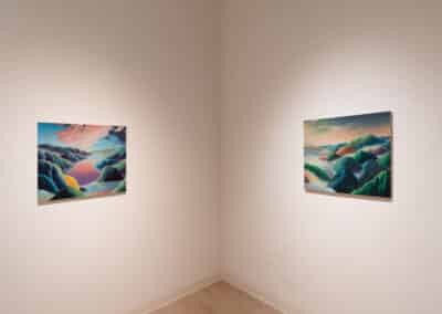 A corner gallery wall displaying two landscape paintings. The painting on the left features colorful, abstract hills and a cloudy sky. The painting on the right depicts a similar scene with vibrant green ridges. Both are illuminated by soft, ambient lighting.