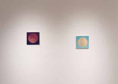 A minimalistic art gallery wall features two square paintings of the moon. The painting on the left has a dark background with a reddish moon, while the one on the right has a lighter background with a yellowish moon. Both are evenly spaced and centered.