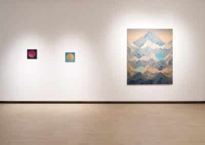 A minimalist art gallery features three pieces: a large painting on the right depicting a range of blue-tinted mountains, and two smaller square paintings on the left, one featuring a red circle and the other a yellow circle, both on dark backgrounds.