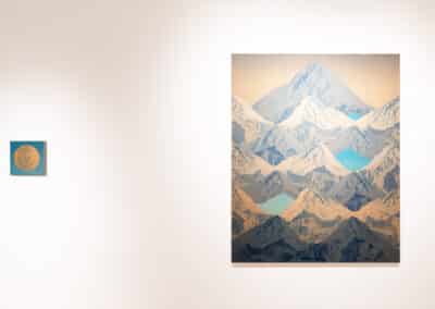 A minimalist art gallery features a large, detailed painting of a mountain range with snowy peaks on a white wall. To its left, a small, circular artwork in blue and beige hues hangs separately. The floor is light-colored, and the ambiance is serene and uncluttered.