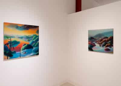 Two colorful landscape paintings hang on a white gallery wall. The left painting depicts a vibrant, surreal sunset over mountains and water, while the right painting shows a serene scene with soft pastel hues and rocky terrain. Both artworks convey a sense of tranquility.