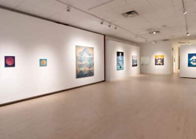 A spacious art gallery displaying mountain-themed artworks on white walls. Paintings of various sizes, primarily featuring mountains and natural landscapes, are evenly spaced along the walls. The floor is light-colored wood, and overhead lighting illuminates the artwork.