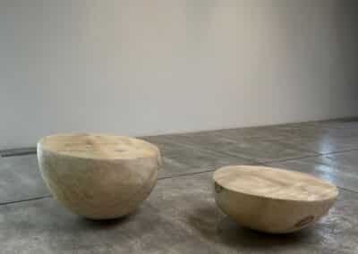 Two wooden hemispherical seating structures crafted by Munson Hunt sit on a polished concrete floor in a minimalist setting. One upright, resembling a bowl, and the other inverted, creating a smooth, flat surface. A plain white wall serves as the background.
